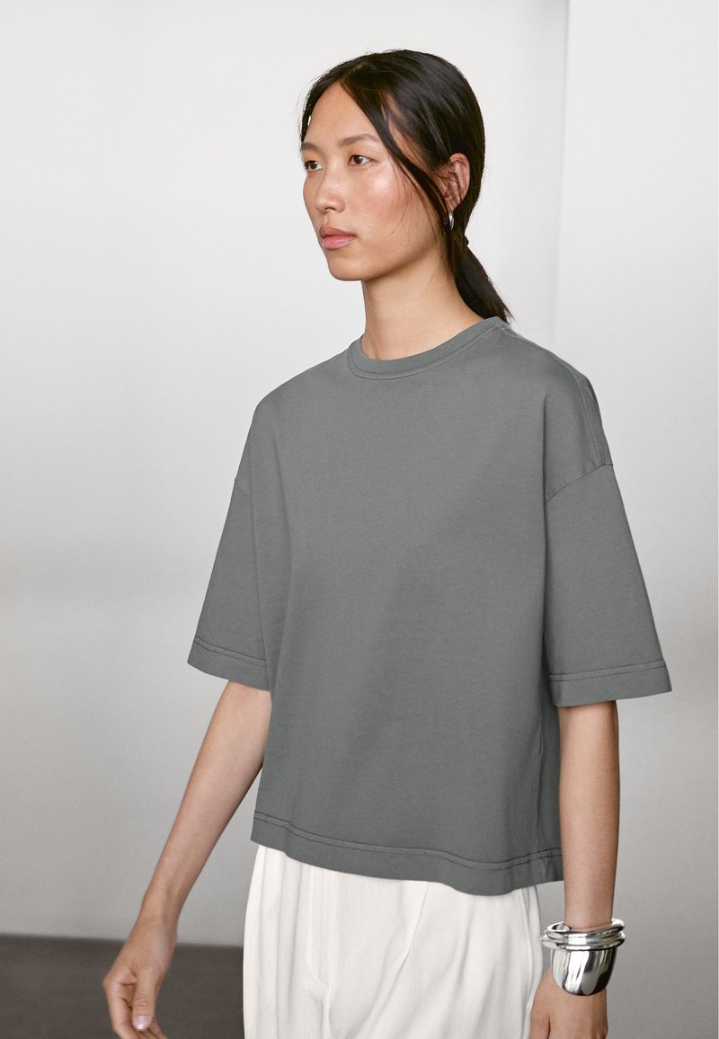 Grå oversized T-shirt lavet af bomuld, med korte ærmer, rund halsudskæring og lige kant. Modellen bærer sølvarmbånd på håndleddet.