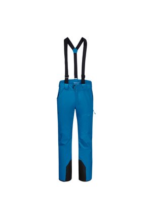 Homme portant un pantalon de ski bleu, des chaussures de ski rouges, un haut noir et des bretelles, tenant des bâtons de ski rouges et noirs en marchant.