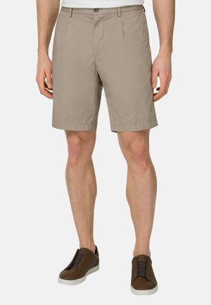 Homme portant un short beige jusqu'aux genoux, une chemise blanche et des baskets marron décontractées, debout devant un fond uni.