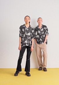 Camicie a maniche corte con stampa grafica in bianco e nero, dalla vestibilità ampia, abbinate a pantaloni neri svasati e pantaloni beige, su uno sfondo chiaro.