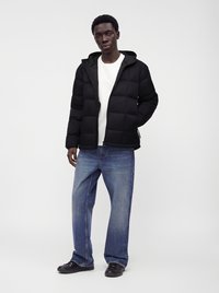 Homme portant une doudoune noire, une chemise blanche, un jean bleu ample et des chaussures noires, debout sur un fond clair uni.