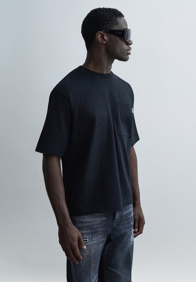 Μαύρο υπερμέγεθες Τ-shirt με κοντά μανίκια και τσέπη στο στήθος. Φορεμένο με ξεβαμμένα ανοιχτόχρωμα τζιν. Σκουρόχρωμα γυαλιά ηλίου ολοκληρώνουν την εμφάνιση.