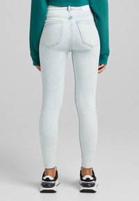 Osoba oblečená v světlých modrých skinny džínách a černobílých teniskách, stojí čelem k jednoduchému šedému pozadí.