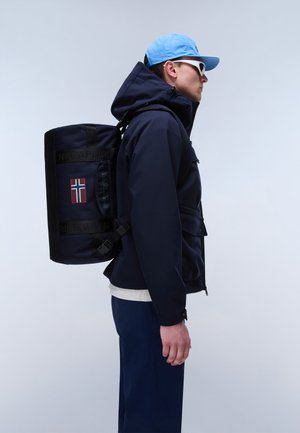 Ein Mann trägt eine marineblaue Jacke, eine blaue Kappe, weiße Sonnenbrillen und einen marineblauen, zylindrischen Rucksack mit einem norwegischen Flaggenaufnäher.