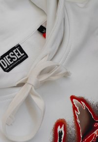 Felpa bianca in tessuto Diesel con cappuccio e coulisse legata a fiocco, etichetta del marchio nera visibile e un design grafico rosso e nero sul tessuto.