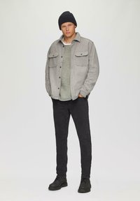 Grau strukturierte Jacke mit zwei Brusttaschen, über einem grünen Pullover getragen, kombiniert mit schwarzen Jeans und schwarzen Schnürstiefeln. Schwarze Strickmütze.