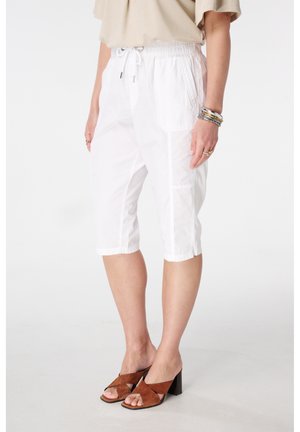 Femme portant un pantalon blanc à cordon jusqu'aux genoux, un haut beige, des sandales à talons marron, et plusieurs bracelets au poignet gauche.