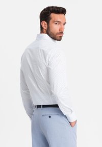 Camicia bianca con bottoni, caratterizzata da piccoli puntini blu, indossata con pantaloni azzurri chiari. La camicia ha un colletto classico e maniche lunghe.