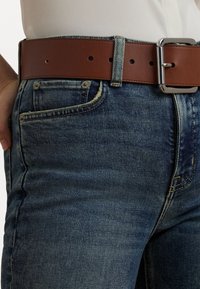 Brun lærreim med en sølvspenne, båret over blå denimjeans med en falmet vask og synlige sømdetaljer.