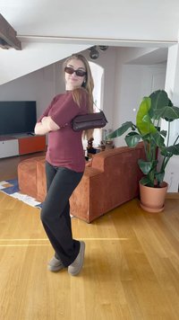 Mulher vestindo uma blusa canelada bordô e calças pretas, segurando uma pequena bolsa castanha. O fundo inclui uma planta verde e um sofá redondo e texturizado.
