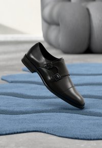 Μαύρο δερμάτινο παπούτσι με διπλούς αγκράφες, μυτερή μύτη, λεία υφή, ανυψωμένη σόλα, τοποθετημένο πάνω σε μπλε υφασμάτινο χαλί.