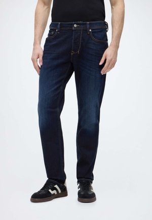 Homme portant un jean bleu foncé et des baskets noires avec des bandes blanches, debout devant un fond uni.