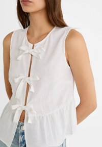 Biała bluzka bez rękawów z trzema kokardkami z przodu, o miękkiej teksturze i marszczonym dołem. Posiada okrągły dekolt i prosty krój.