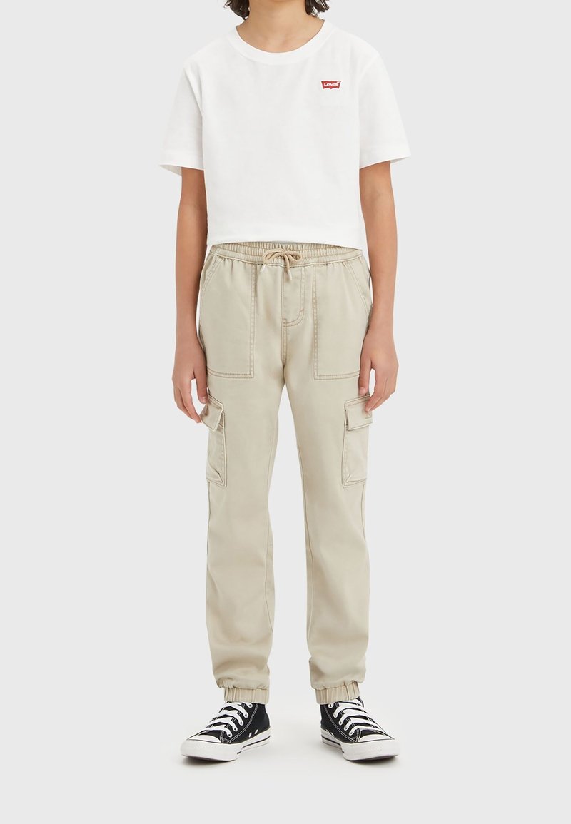 T-shirt blanc raccourci avec logo Levi's rouge, pantalon cargo beige avec poches latérales, taille élastique, et baskets basses noires.