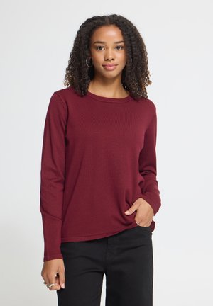 Jeune femme aux cheveux bouclés portant des boucles d'oreilles créoles, un haut bordeaux à manches longues et un pantalon noir, avec une main dans la poche.