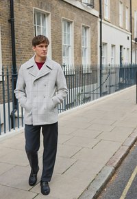 Harmaa ruudullinen kaksirivinen takki, jossa on leveä kaulus ja etutaskut, yhdistetty tummiin housuihin ja mustiin liukkailla kengillä. Kävelee asfaltilla.