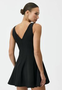 Kobieta z włosami upiętymi w kok, ubrana w czarną, bezrękawnikową sukienkę z głębokim dekoltem na plecach, patrząca przez ramię na jednolitym białym tle.