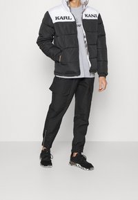 Czarna i biała kurtka puchowa z logo "KARL", szary t-shirt, czarne spodnie cargo i czarne sneakersy z wzorzystą podeszwą.
