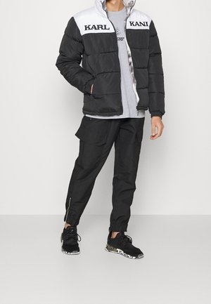 Giacca a vento nera e bianca con logo "KARL", t-shirt grigia, pantaloni cargo neri e sneakers nere con suola decorata.