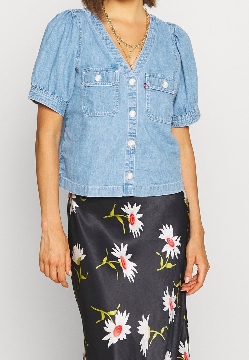 Chemisier cropped en denim bleu clair avec manches bouffantes, col en V, orné de deux poches poitrine et de boutons blancs. Associé à une jupe fleurie.