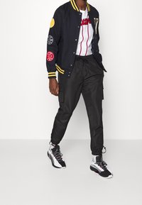 Man draagt een zwart varsityjack met emblemen, wit honkbalshirt, zwarte cargobroek en zwart-witte sneakers, staand tegen een witte achtergrond.