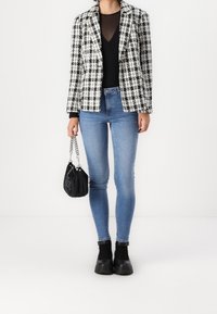 Blazer a cuadros en blanco y negro sobre una blusa negra, combinado con jeans ajustados de color azul claro y botines de plataforma negros, sosteniendo un bolso negro.