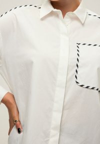 Weißes Hemd mit schwarzem Streifenakzent an den Schultern und der Tasche. Hergestellt aus glattem Stoff, bietet es ein lockeres Design und einen entspannten Schnitt.