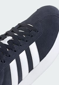 Granatowe zamszowe sneakersy z białymi skórzanymi paskami, płaskimi czarnymi sznurówkami i fakturowaną białą gumową podeszwą, prezentujące klasyczny sportowy design.