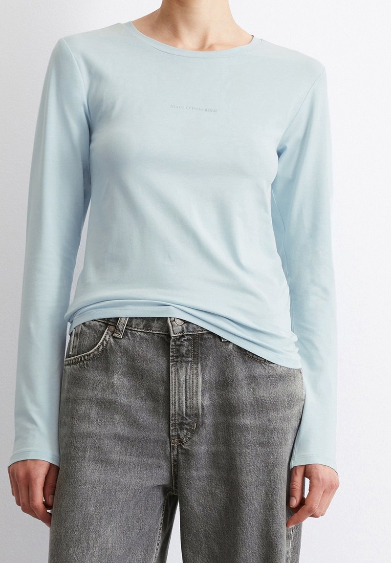 Vaaleansininen pitkähihainen paita, valmistettu pehmeästä kankaasta, jossa pyöreä pääntie ja hienovarainen logoteksti. Yhdistetty haalistuneisiin harmaisiin farkkuihin.