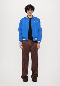 Cazadora bomber de satén azul con acentos amarillos, camiseta negra, pantalones anchos de satén marrón y zapatos negros tipo slip-on. Gafas de sol rectangulares y transparentes.