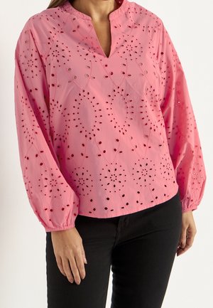 Vrouw met een roze blouse met lange mouwen en geborduurde bloemmotieven, en een zwarte broek, afgebeeld van schouders tot heupen.