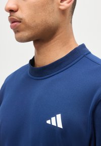 Νέος άνδρας που φοράει μπλε φούτερ με μικρό λευκό γεωμετρικό λογότυπο στο αριστερό στήθος, εμφανιζόμενος από τους ώμους και πάνω, στραμμένος προς τα αριστερά.