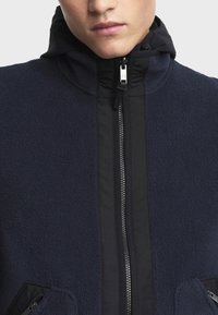 Mornarica flis jakna s visokim ovratnikom, prednjim patentnim zatvaračem i dva bočna džepa; sadrži glatku, kontrastnu crnu tkaninu uz patent zatvarač.