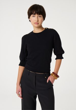 Schwarzer Strickpullover mit Puffärmeln und Rüschensäumen. Ausgestattet mit goldfarbenen Knöpfen an der linken Schulter. Getragen mit schwarzen Hosen und Armreifen.