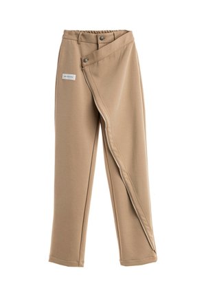 Joven con el pelo trenzado que lleva una chaqueta beige oversize con grandes bolsillos, pantalones a juego y zapatos blancos gruesos, de pie frente a un fondo blanco.