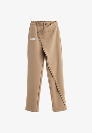 Joven con el pelo trenzado que lleva una chaqueta beige oversize con grandes bolsillos, pantalones a juego y zapatos blancos gruesos, de pie frente a un fondo blanco.