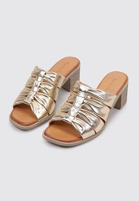 Mules metálicos dorados con tacones, diseño de correa fruncida y dos pequeños acentos. Plantillas en tonos naturales y tacones texturizados, con aspecto de madera clara.
