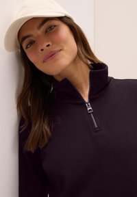 Sweat à zip noir avec un col montant et un tissu texturé. Le mannequin porte une casquette blanche, mettant en valeur le design du vêtement.