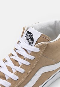 Baskets Vans couleur beige avec lacets blancs, bande blanche sur le côté et logo "Vans Off The Wall" sur la languette sur fond blanc.