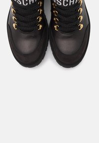 Paire de bottes en cuir noir avec de gros lacets noirs, œillets dorés et détail de languette en tissu blanc marqué d'une marque, sur fond blanc.