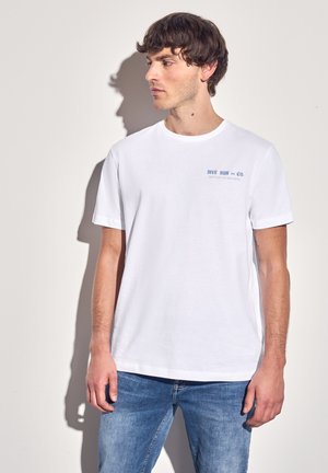Witte katoenen t-shirt met korte mouwen, ronde halslijn en blauwe opdruk "DIVE SUN and CO." op de linkerborst. Draagt een blauwe spijkerbroek.