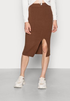 Gonna midi in knit marrone con uno spacco laterale, caratterizzata da una vita a coste. Abbinata a sneaker beige, su uno sfondo bianco uniforme.