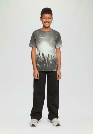 Grijze t-shirt met korte mouwen met een grafisch ontwerp van opgestoken handen, gecombineerd met zwarte wijde broek en lichtgekleurde sneakers.