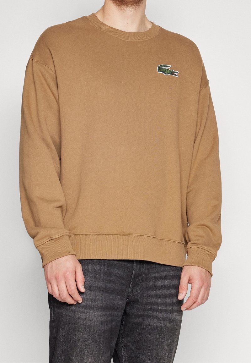 Sweat-shirt marron en tissu doux avec un col rond et des manches longues. Il présente un logo vert brodé sur le côté gauche de la poitrine.