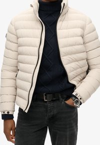 Giacca puffa di un beige chiaro, con chiusura zip, dettagli neri e cuciture orizzontali. Indossata sopra un maglione lavorato a maglia blu navy e jeans neri.