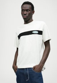 Svijetlosiva pamučna majica s horizontalnom crnom prugom i plavom oznakom na prednjoj strani, kratkih rukava i opuštenog kroja.