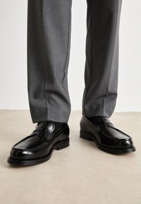 Melnas ādas loafers ar gludu apdari, ar noapaļotu purngalu un dekoratīvu jostu, kopā ar pelēkiem biksēm.