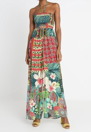Femme portant une robe maxi bustier à col halter avec des motifs floraux et géométriques colorés et des sandales jaunes à talon bloc.