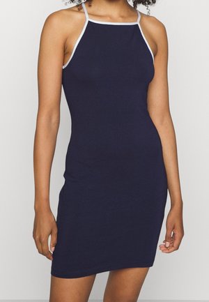 Femme portant une robe sans manches bleu marine ajustée avec un liseré blanc autour de l'encolure et des bretelles, debout devant un fond uni.