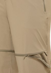 Pantaloni leggeri beige con una cerniera orizzontale parzialmente aperta sopra il ginocchio, che mostra un design delle gambe convertibile.